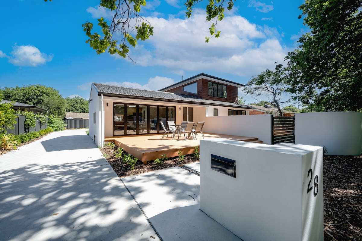 The exterior of a modern family home is depicted, featuring a spacious wooden deck with outdoor seating. The driveway leads to the entrance, accented by landscaped greenery. A white mailbox stands prominently by the paved walkway, with a backdrop of trees and a clear blue sky.