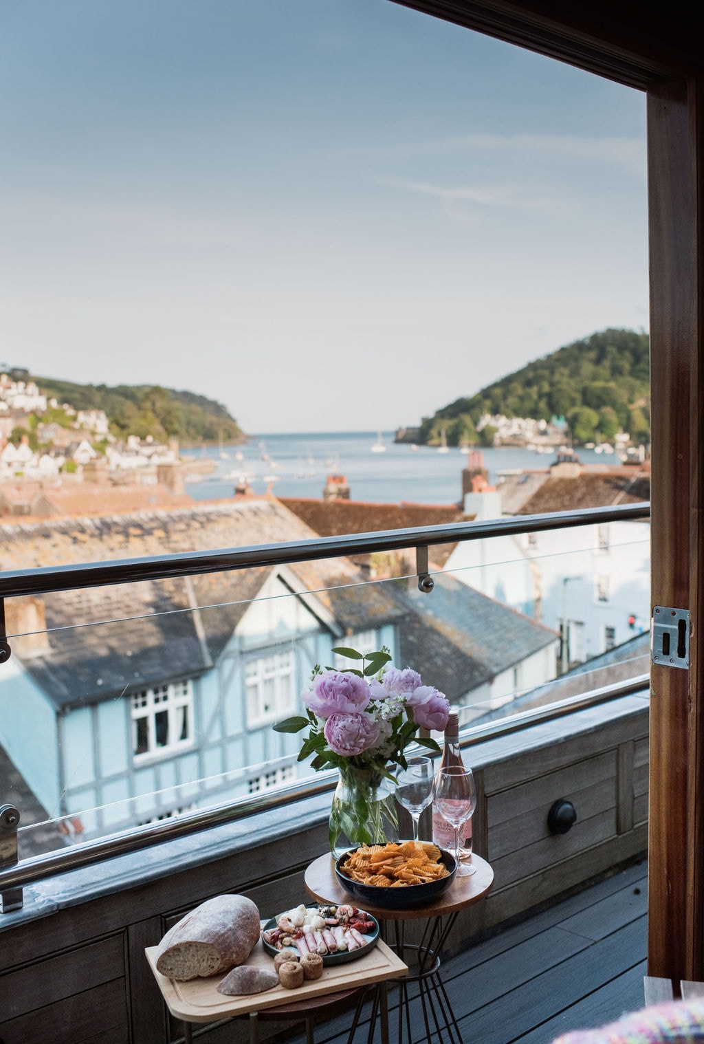 A balcony is shown with a view of the River Dart and surrounding hills. A small table displays a bouquet of flowers, a bottle of rose, and plates of food. The scene captures serene waters, quaint houses, and a clear sky.