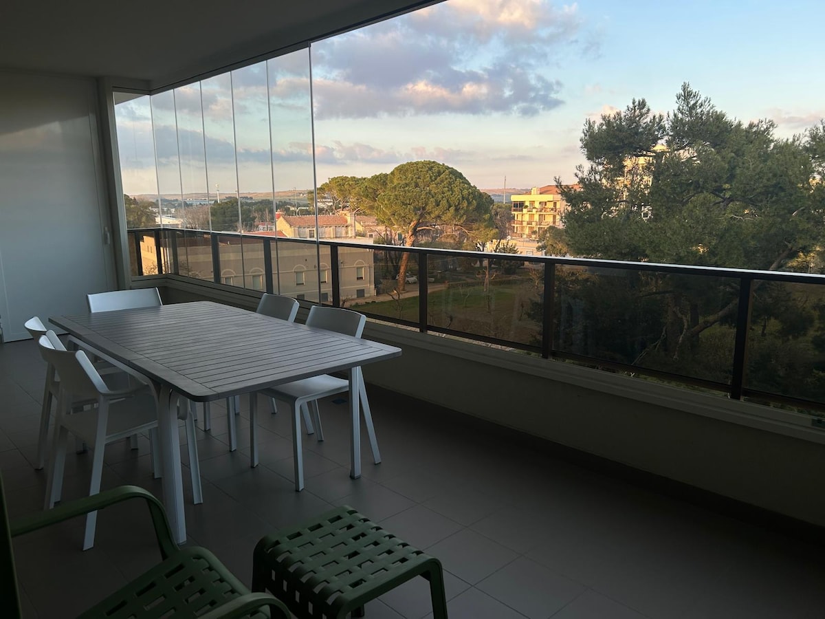 A spacious terrace is featured, furnished with a dining table and several white chairs, overlooking a tranquil residential area. Large glass panels offer unobstructed views of the surrounding greenery and distant buildings, with soft natural light illuminating the space.