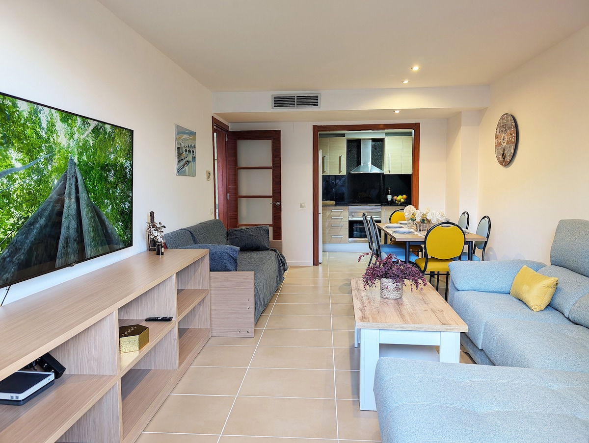 A spacious living area features a large Smart TV mounted on the wall. Comfortable seating arrangements include a light grey sofa and a cozy chair. A dining table with chairs is seen in the background, with an open kitchen equipped for cooking visible through an adjoining entry.
