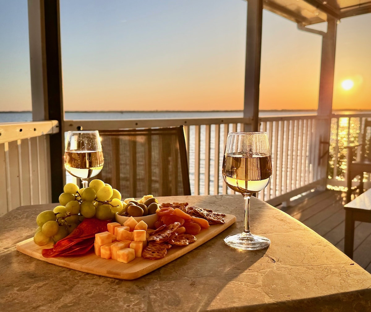 A table is set on a covered deck with two glasses of white wine beside a wooden cheese board. The board features an assortment of grapes, assorted cheeses, olives, and cured meats. A vibrant sunset casts a warm glow over the water in the background.