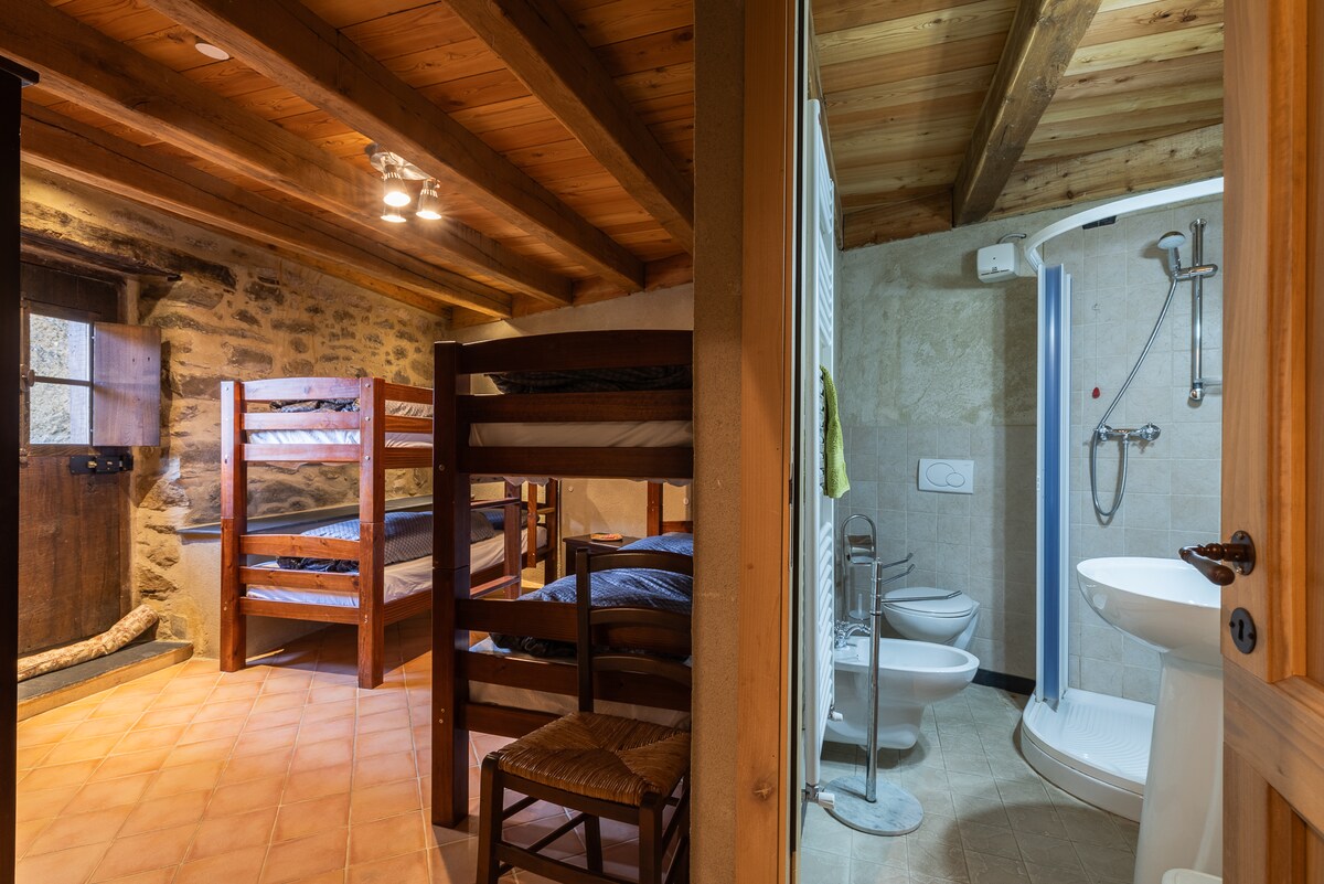 A rustic sleeping area features wooden bunk beds against a stone wall, complemented by a soft earth-toned tiled floor. An adjoining bathroom includes a shower, toilet, and sink, all set within a modern design. Wooden beams add a cozy touch to the ceiling.