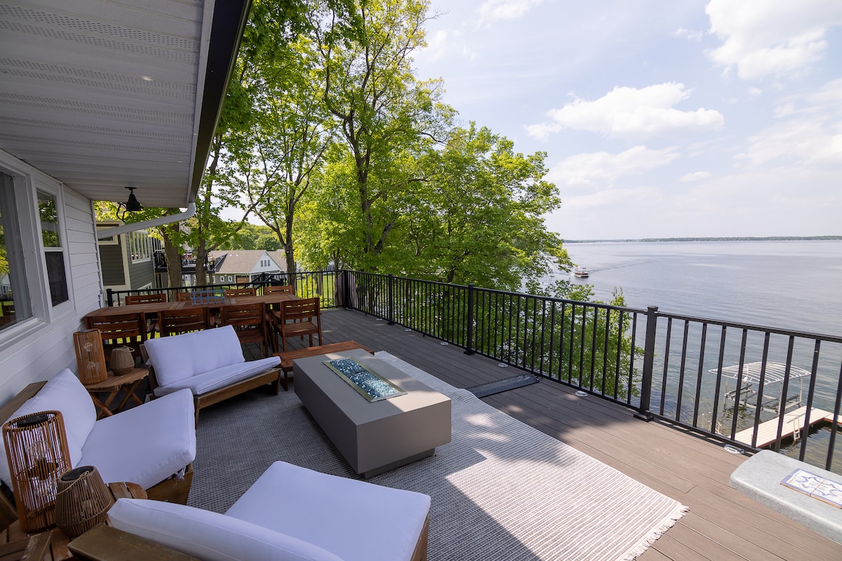 A spacious deck overlooking the water features comfortable seating arrangements, including a modern sectional couch and a soft rug. A gas fire table is positioned at the center, offering views of the serene lake and surrounding greenery.