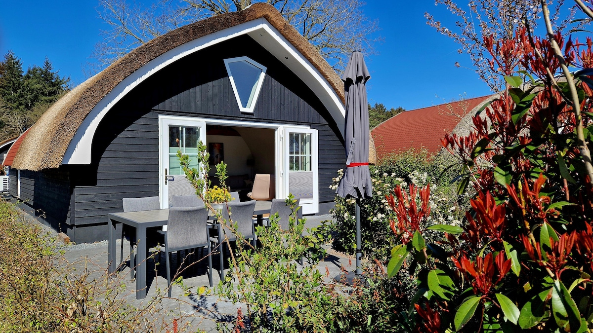 The exterior of a charming house is viewed, featuring a thatched roof and a triangular window. A shaded patio area with a dining table and chairs is surrounded by lush greenery and colorful flowers, creating a serene outdoor setting.