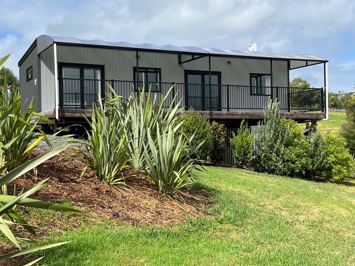 A newly built cottage is set among lush greenery, elevated on a wooden platform. The exterior features a covered porch with railings, large windows, and a modern facade. Surrounding plants and grass create a natural feel, offering a connection to the tranquil environment.