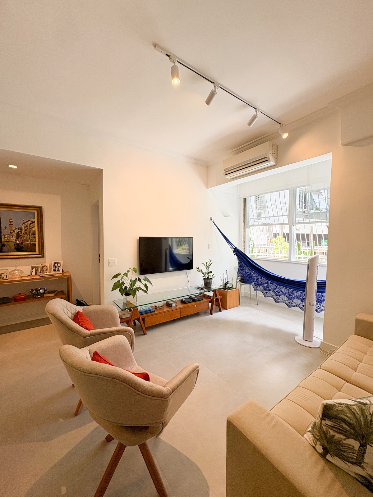 A bright living area features a comfortable sofa and two armchairs with red cushions. A wall-mounted TV is positioned across from the seating. Natural light streams in through large windows, while a hammock is draped in a corner, complemented by indoor plants.
