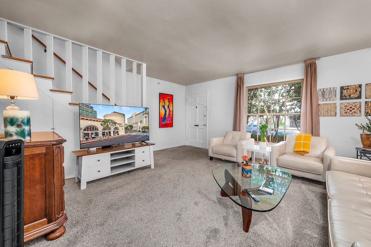 A spacious living room is presented, featuring a large television on a white media console. Two light-colored sofas are arranged around a glass coffee table. Natural light enters through a window, while artwork adds character to the walls. A soft carpet covers the floor.