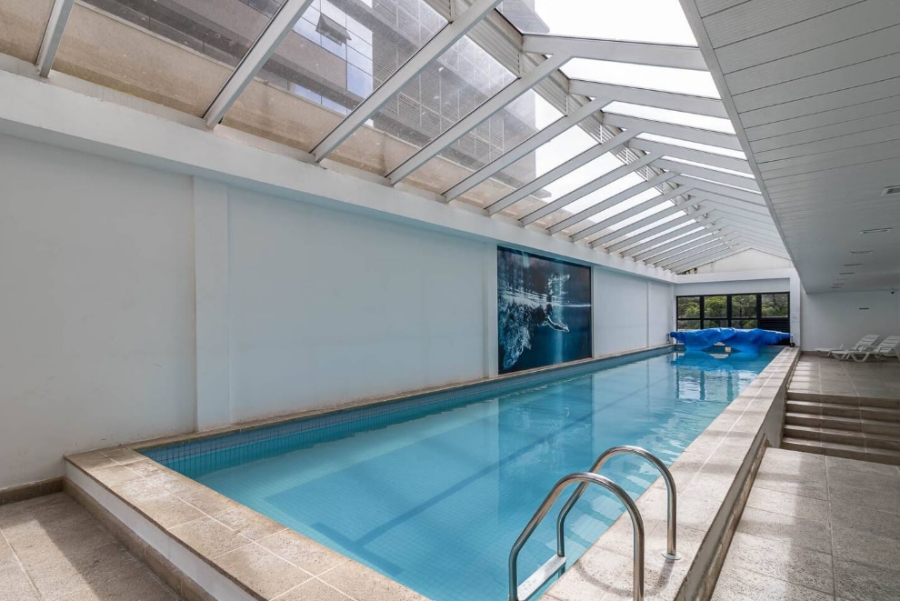 An indoor pool area is highlighted by large windows and a clear roof, allowing natural light to fill the space. The water appears calm and inviting, with a depth marker visible on the poolside. Lounge chairs line the edge for relaxation.