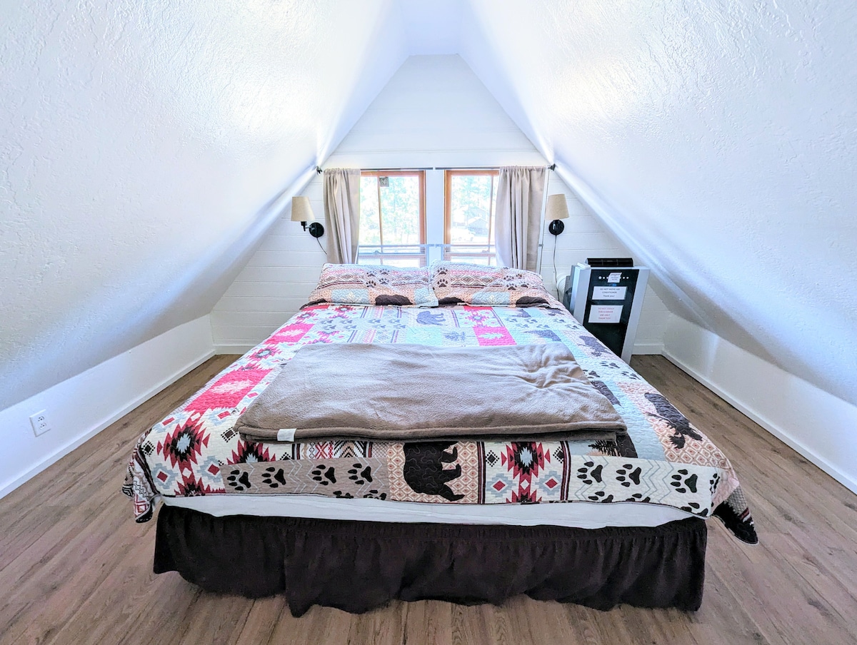 A cozy bedroom is presented under a sloped ceiling, featuring a spacious bed adorned with patterned bedding. Two bedside lamps are placed on either side, and a small nightstand offers storage. Natural light filters in through the large windows, enhancing the welcoming ambiance.