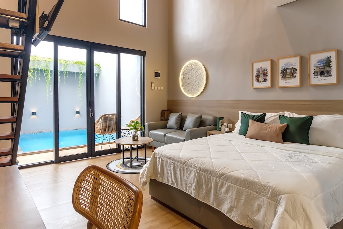 A spacious living area features a comfortable bed with soft linens and accent pillows. A seating arrangement, comprising a sofa and two chairs, faces large glass doors that open to a private pool. Natural light fills the room, enhancing the serene atmosphere.