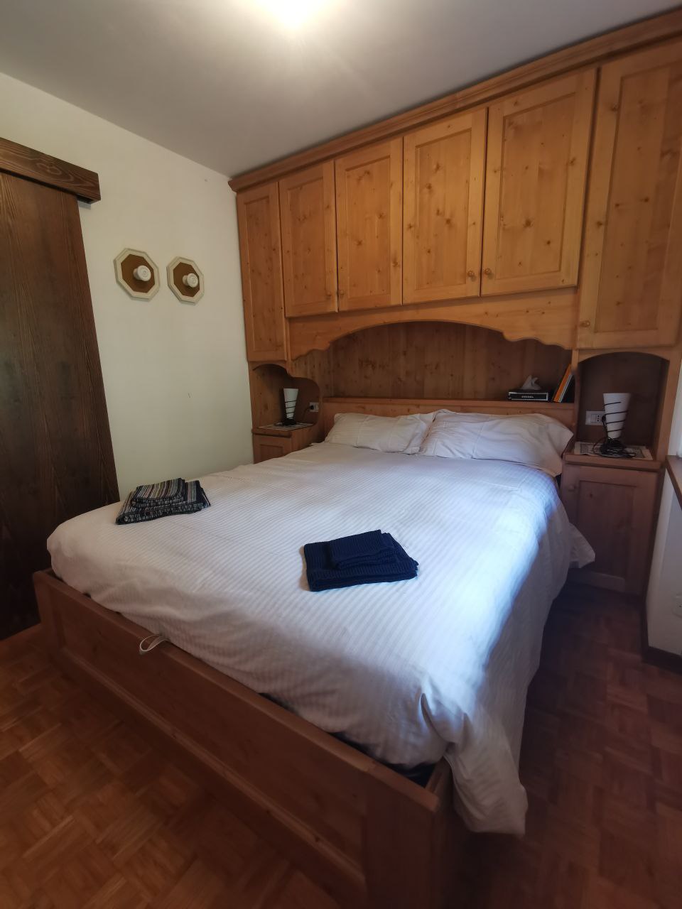 A spacious bedroom featuring a wooden bed frame with a neatly made bed and coordinated linens. A wooden headboard with built-in storage complements the room. Two decorative plates are displayed on the wall, and surrounding wooden cabinets add to the rustic charm.