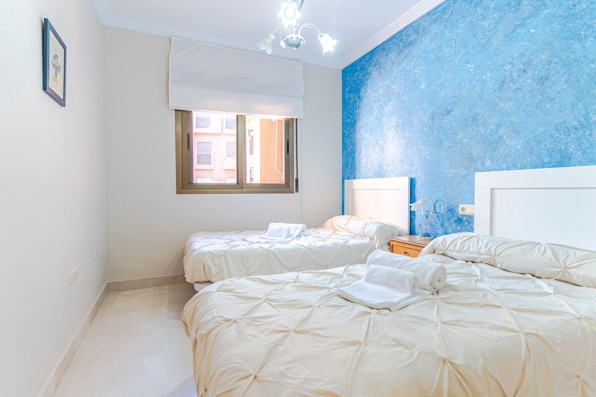 غرفة نوم مع سريرين مفردين، وألواح رأس بيضاء، وبياضات سرير خفيفة ومناشف، ومنضدة خشبية، وجدار أزرق مزخرف، وخزانة مدمجة، ونافذة مع مصراع والوصول إلى الحمام، على أرضية بلاط خفيفة.
