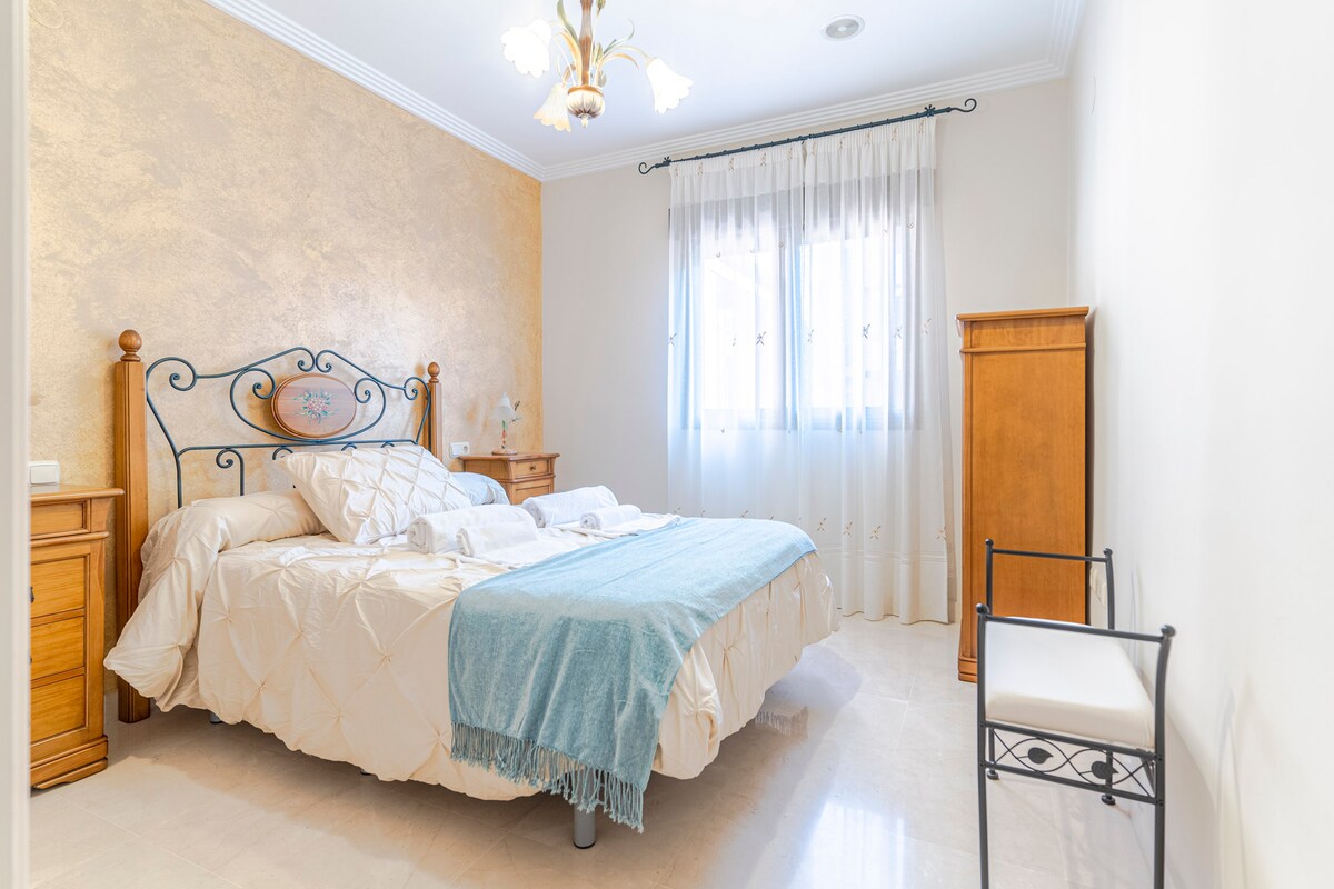 غرفة نوم بسرير مزدوج، ولوح سرير أمامي معدني، وأغطية سرير بيضاء وبطانية زرقاء، وطاولات خشبية وخزانة، وخزانة ملابس مدمجة، وكرسي جانبي، ونافذة بستائر بيضاء، وجدران بيضاء ودرجات لونية خوخية، وأرضيات من الخشب الفاتح.
