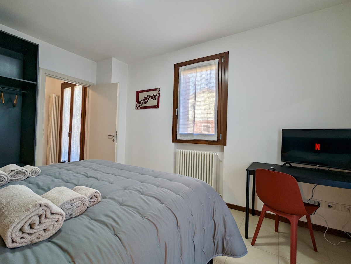 A spacious bedroom features a large bed with neatly arranged towels, complemented by a desk and chair. Natural light enters through a window with light curtains, and a minimalist wardrobe provides storage space. A door leads to a private bathroom.