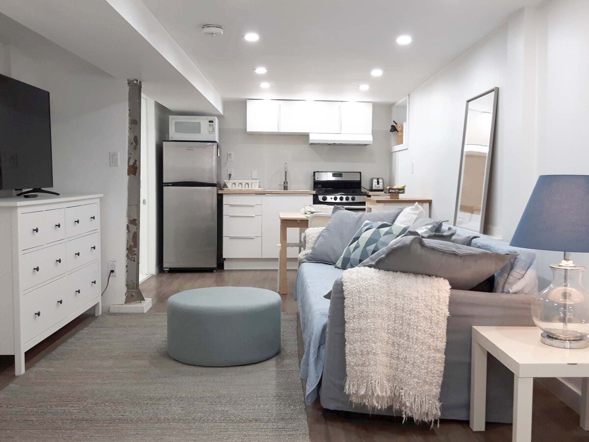 An open-concept studio features modern appliances and clean lines. A cozy seating area with a light gray sofa is complemented by a round ottoman. Natural light is provided by overhead fixtures and a nearby window, while a neutral color palette contributes to the serene ambiance.