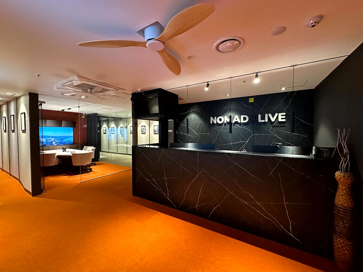The reception area features a modern design with a dark marble front desk labeled 'NOMAD LIVE.' Soft lighting illuminates the space, highlighting an orange carpet and contemporary decor. A separate seating area with glass walls is visible in the background, accommodating small group discussions.