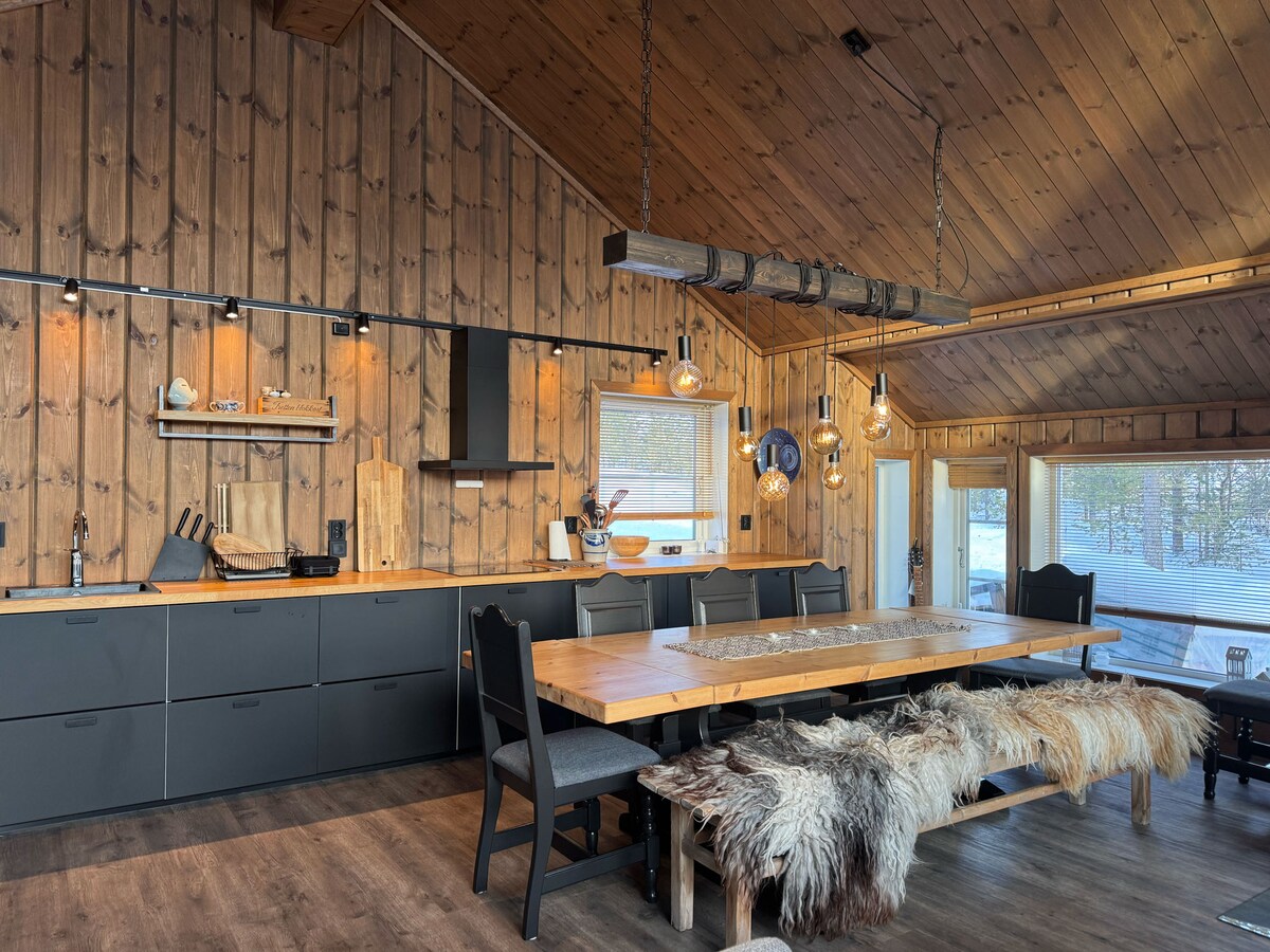 A spacious kitchen with wooden walls and high ceilings features a long dining table that accommodates multiple guests. Modern kitchen appliances and ample storage are visible. Pendant lighting adds warmth, enhancing the inviting atmosphere. Large windows offer views of the surroundings.