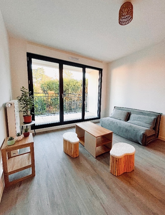 A luminous living area is captured, featuring a convertible sofa and a wooden coffee table. Two textured stools are positioned beside the table. Large glass doors lead to a balcony, allowing natural light to fill the room and offering views of greenery outside.