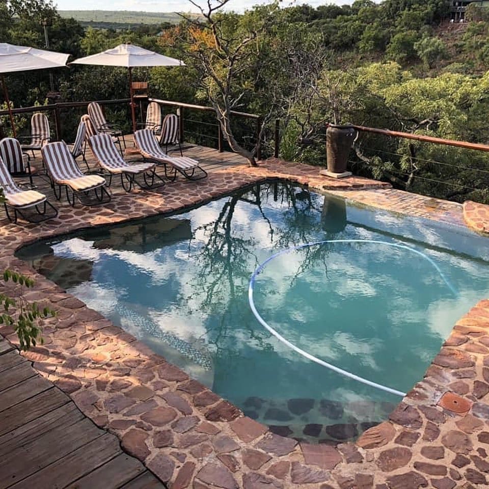 A rectangular pool is surrounded by a stone deck featuring lounge chairs with striped fabric. Umbrellas provide shade nearby, and the water reflects the surrounding trees and sky. The natural landscape creates a serene atmosphere.