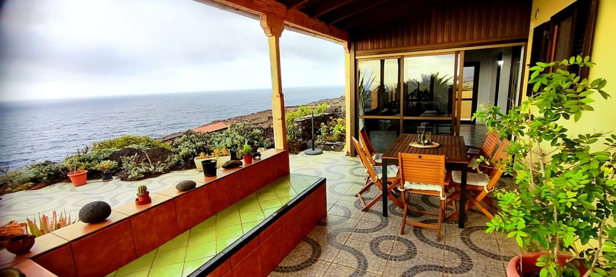 An outdoor terrace is visible, featuring wooden dining furniture, surrounded by lush greenery and potted plants. A panoramic view of the ocean extends in the background, with the sun casting a soft light over the space. Textured tiles add character to the area.