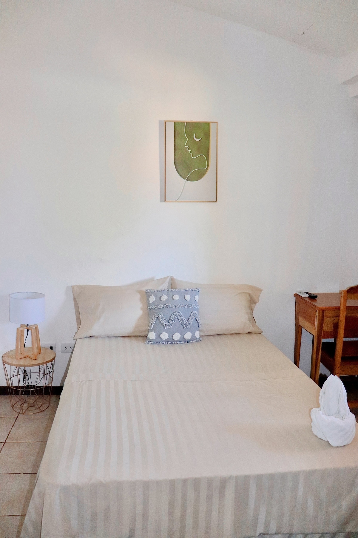A spacious sleeping area offers light-colored bedding with a decorative pillow featuring a seashell design. A small lamp is positioned next to the bed on a wooden table. A wooden chair and desk are visible in the background, complemented by a minimalistic wall art piece.