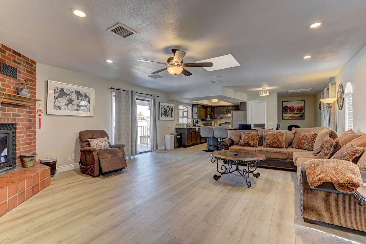 An open living space features a comfortable seating area with a sectional sofa and a recliner chair. Natural light enters through large windows, highlighting the wood flooring. A cozy fireplace with a brick surround adds warmth to the room, which connects to a visible kitchen area.