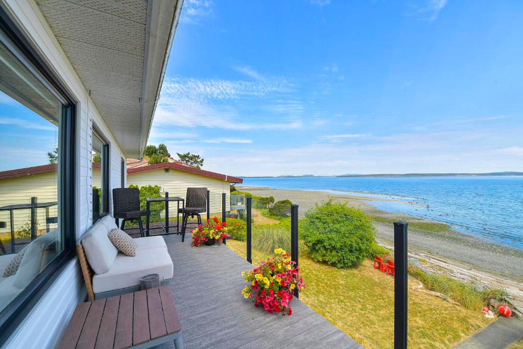 A spacious deck overlooks the ocean, featuring comfortable seating and vibrant flower pots. The serene water stretches out toward the horizon, with gentle waves lapping against the shore. The surrounding landscape includes greenery and a gravel beach, inviting a peaceful outdoor experience.
