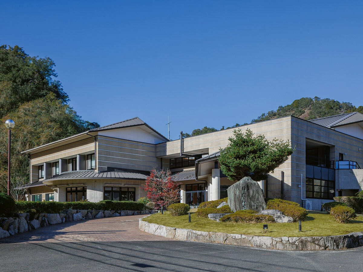 The exterior of a modern building is set against a clear blue sky, featuring clean lines and natural stone elements. Seasonal foliage frames the entrance, and well-maintained landscaping enhances the inviting ambiance of the property.