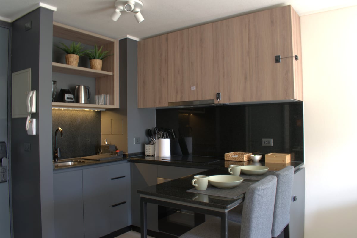 A modern kitchen area is equipped with stainless steel appliances and ample storage. Countertops in dark tones complement the sleek cabinetry. A small dining table is set with two plates and utensils, surrounded by comfortable seating, creating an inviting space for meals.