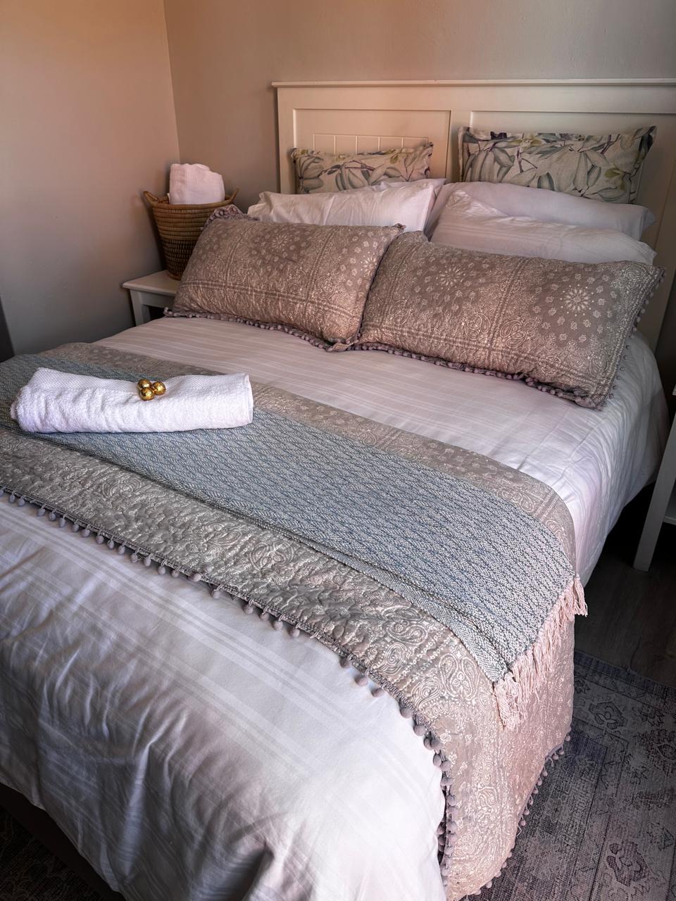 A well-appointed sleeping area features a spacious bed topped with textured pillows and a decorative throw. The neutral colour palette is complemented by subtle patterns. A set of fresh towels is neatly arranged beside a woven basket on the side table.