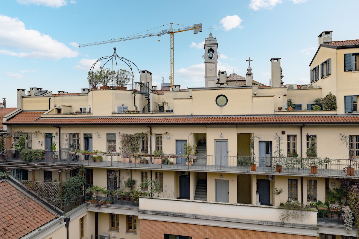 A view of a charming building featuring multiple balconies adorned with potted plants. The architectural details include a dome and a clock tower in the background, with a clear sky highlighting the structure's inviting ambiance.