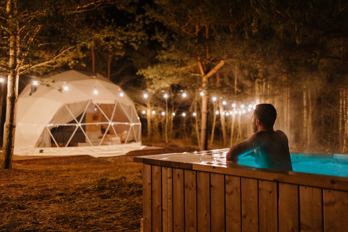 A private wooden hot tub is shown, with a person relaxing in the warm water. In the background, a dome-shaped glamping structure is visible, illuminated by string lights that enhance the serene atmosphere of the forested area.
