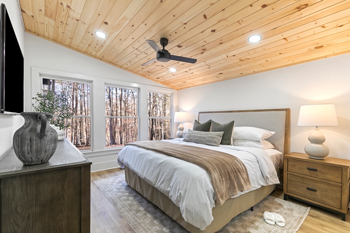 A well-lit bedroom features a spacious bed with neutral bedding and decorative pillows. Two bedside tables with lamps are placed on either side. A modern ceiling fan is overhead. Large windows showcase views of surrounding trees, enhancing the natural light in the room.