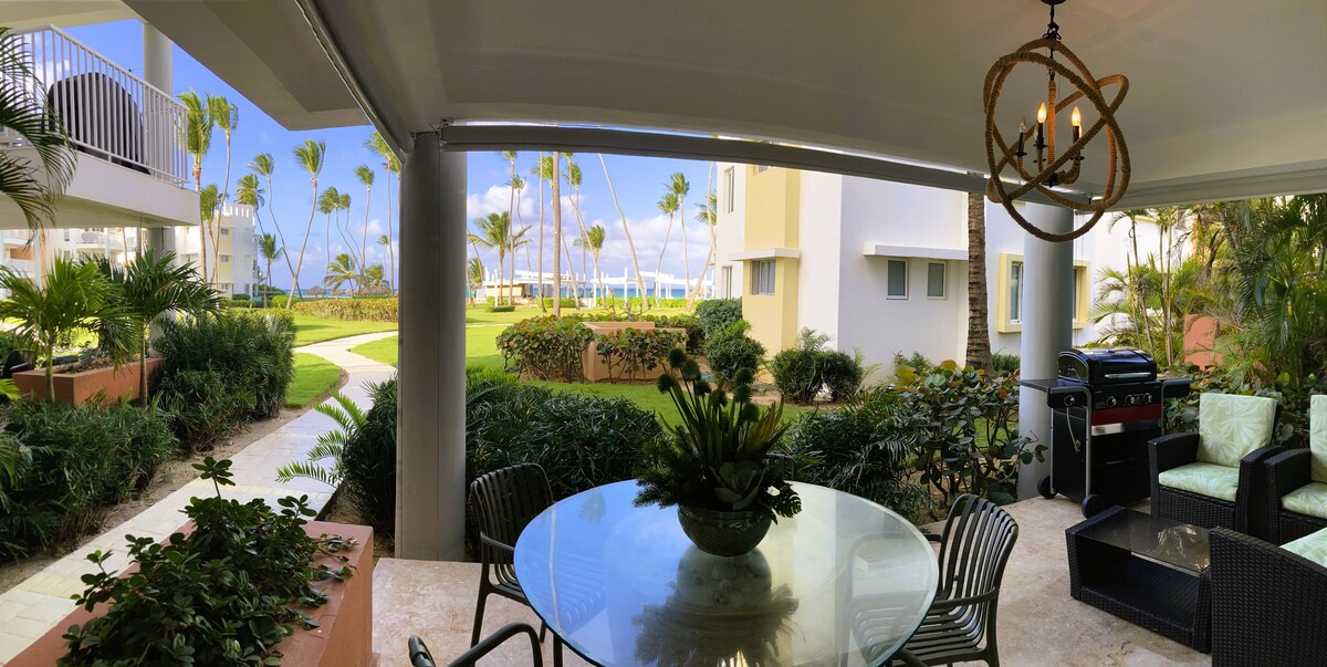 A private terrace features a round dining table surrounded by chairs, overlooking a lush garden pathway leading to the beach. A gas BBQ is positioned nearby, with comfortable seating visible for relaxation. The scene is framed by palm trees and an expansive view of the sea.