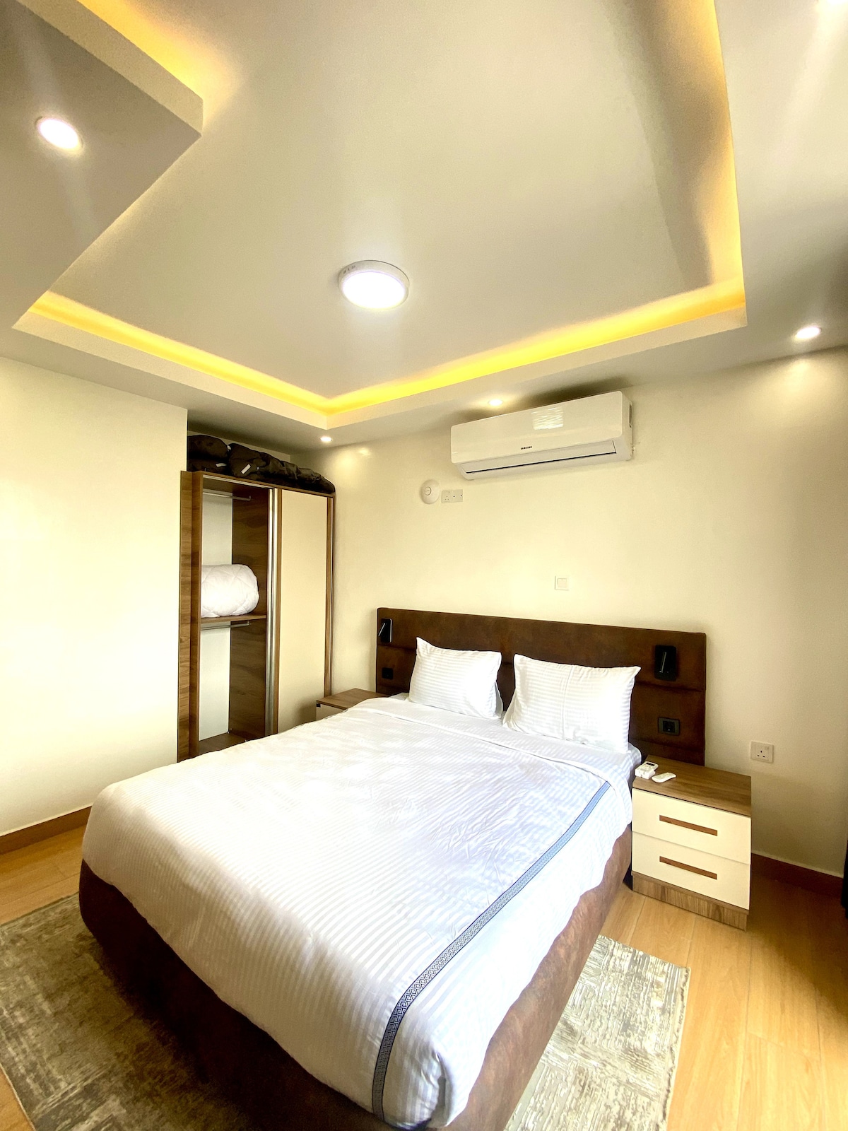 The well-appointed room showcases a plush king-sized bed with crisp white linens and two comfortable pillows. A modern wardrobe is positioned against one wall, while soft lighting emanates from the ceiling, creating a warm ambiance. An air conditioning unit is visible for climate control.