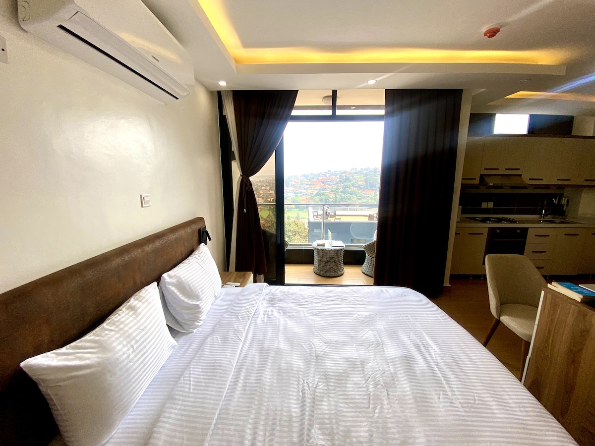 A cozy sleeping area features a plush king-sized bed dressed in white linens. Floor-to-ceiling windows offer a panoramic view of the cityscape, while muted curtains frame the scene. The adjoining kitchenette is visible in the background, providing a well-equipped cooking space.