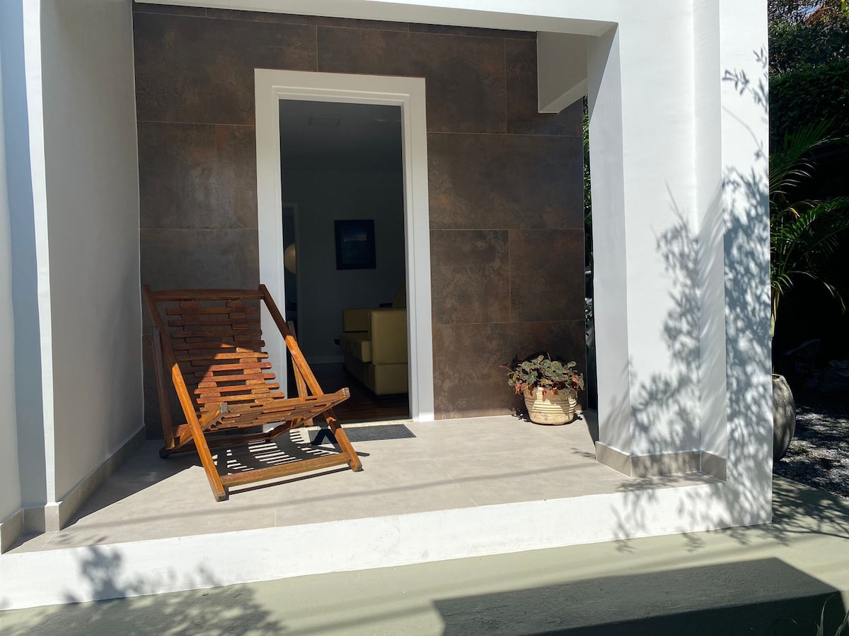 An outdoor area features a wooden lounge chair positioned on a tiled patio. A potted plant adds a touch of greenery next to the entrance, framed by the home's light exterior and dark accent wall. Soft shadows create a calming effect.