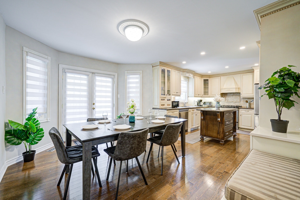 A modern kitchen and dining area is featured, with a dining table set for four. Elegant cabinetry and a spacious island are present, with ample natural light filtering through the window treatments. A small potted plant adds a touch of greenery to the inviting space.