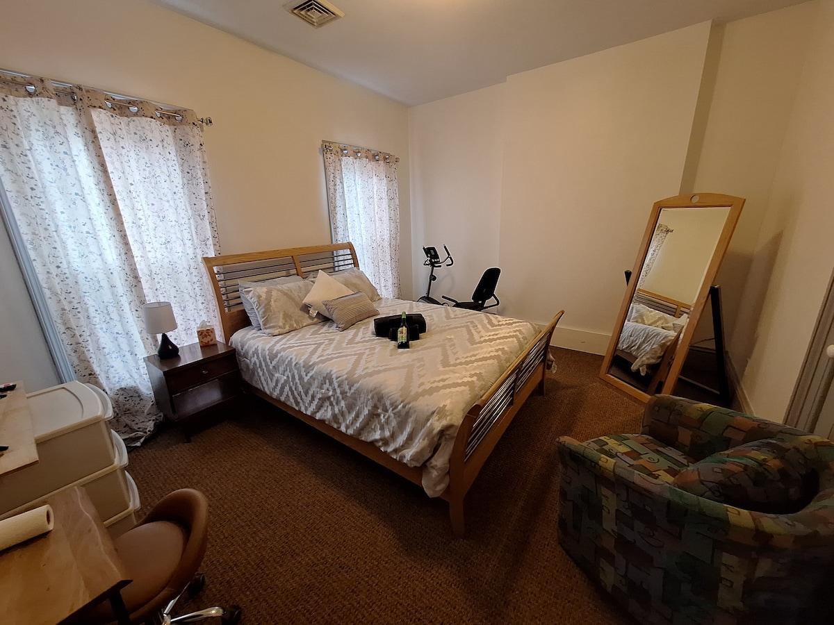 The private room features a full-size bed with a neutral-patterned blanket, flanked by two windows adorned with light curtains. A standing mirror and comfortable chair are positioned beside a nightstand with a lamp. A small desk with a chair provides a suitable workspace.
