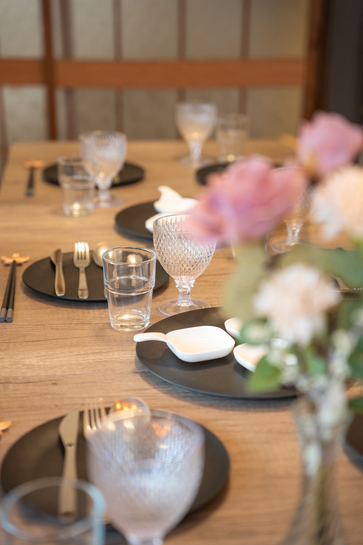A dining table is set with elegant black plates, sparkling glassware, and polished cutlery. Soft focus flowers add a touch of color, while the warm wood grain of the table creates a cozy ambiance.
