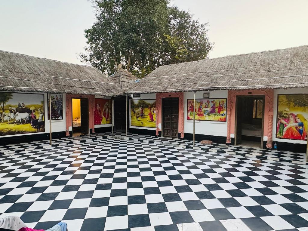 A spacious courtyard features black and white checkered flooring, bordered by straw-roofed structures. Walls are adorned with vibrant murals depicting cultural scenes, contributing to a lively and inviting atmosphere. The surrounding greenery adds a touch of tranquility to the setting.