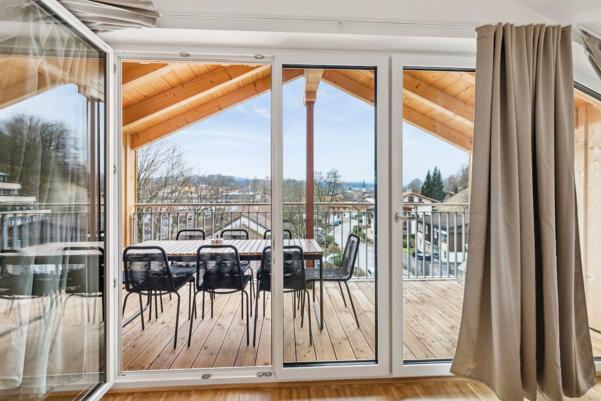 A spacious balcony is visible through large glass doors, showcasing a wooden deck and a dining table with chairs. The sloped wooden ceiling adds a cozy touch. Scenic views of nearby trees and houses can be seen in the background, under a clear sky.
