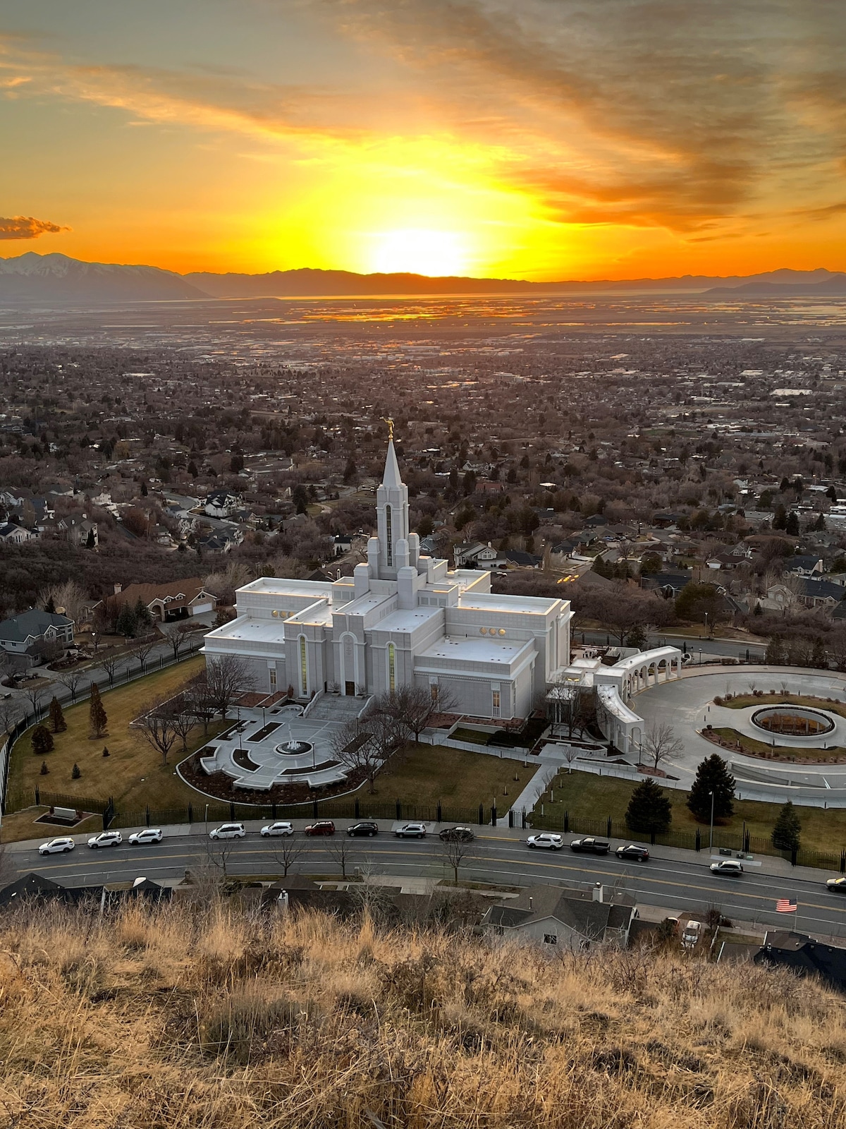 A scenic view captures a temple bathed in the golden glow of sunset, surrounded by a sprawling landscape. Majestic mountains loom in the distance while the structures below reflect the warm hues of the sky, creating a serene and peaceful ambiance.