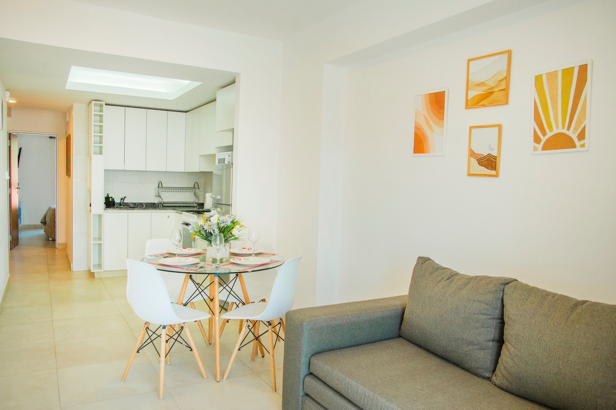 A modern open-concept living space features a cozy gray sofa and a round dining table set for four. Bright artwork adorns the walls, and the kitchen is equipped with essential appliances. Natural light brightens the neutral tones throughout the area.