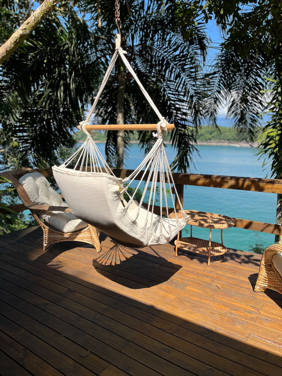 A hammock is gently suspended between trees on a wooden deck, offering a serene view of the calm sea. Nearby, two comfortable chairs and a small side table are positioned on the polished wooden flooring, surrounded by lush greenery.