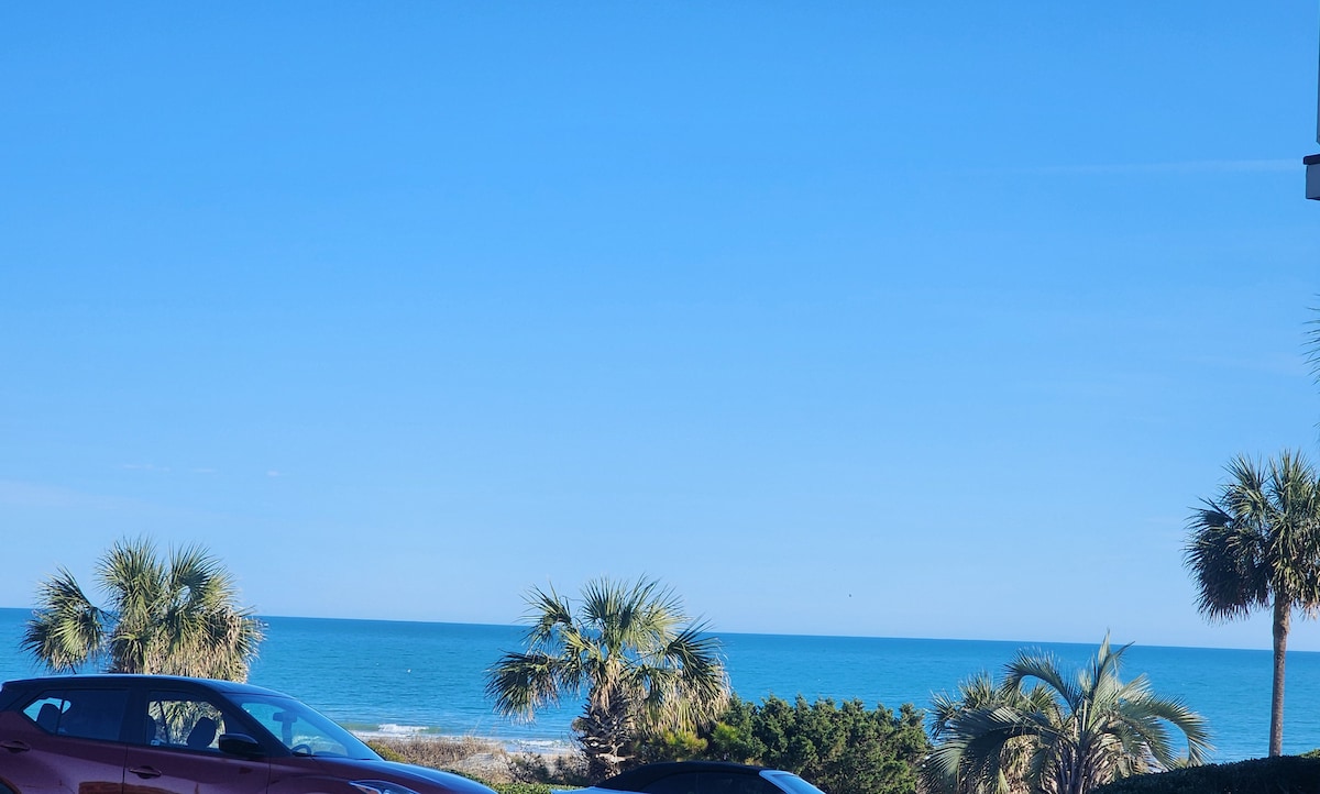 A clear blue sky meets the serene ocean horizon, with gentle waves visible from a distance. Palm trees add a tropical touch in the foreground. Two parked cars can be seen, providing context for the condo's coastal location.