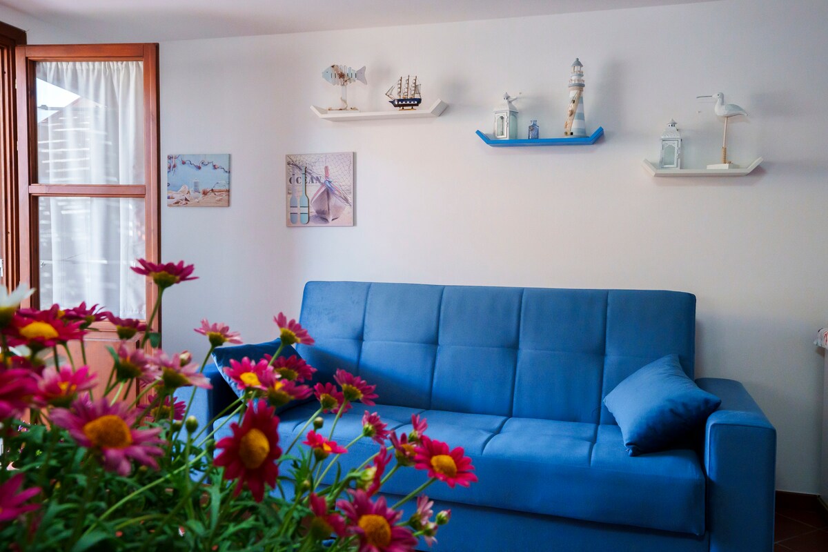 A blue sofa is positioned against a light-colored wall, complemented by nautical-themed decor on shelves above. Framed photographs and small decorative items, including a lighthouse, are displayed. A vase of pink flowers in the foreground adds a touch of color to the inviting living area.