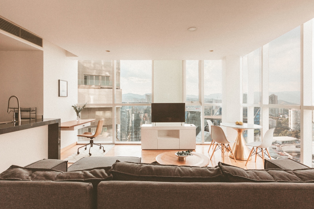 A spacious living area showcases large floor-to-ceiling windows that provide a sweeping view of the city landscape. A modern sectional couch faces a sleek entertainment unit, while a dining area with white chairs and a wooden table is situated nearby. A desk area is also visible.