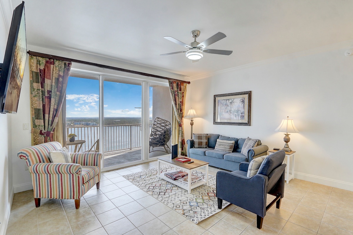 A light-filled living area is presented with a sofa and armchair, complemented by a coffee table on a patterned rug. Large glass doors lead to a balcony, offering views of the surroundings. Decorative lamps add a soft glow, while a ceiling fan enhances comfort.