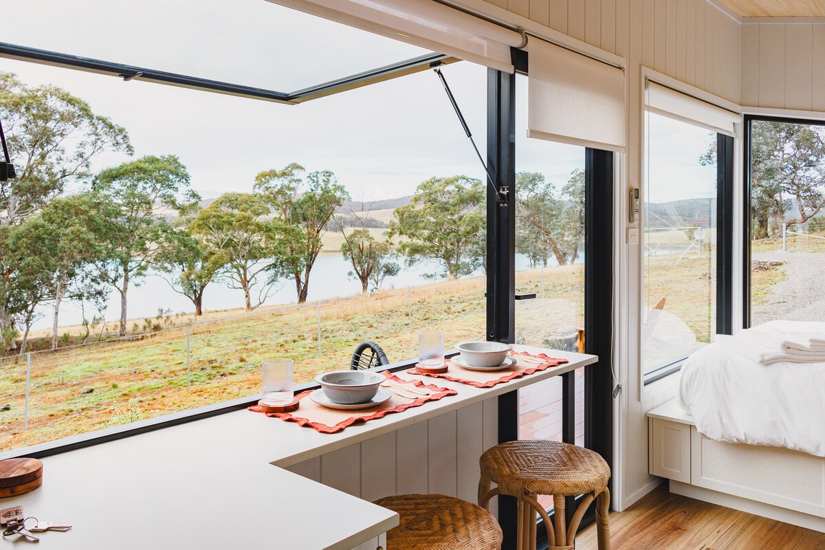 A dining area features a clean countertop with two place settings, including bowls and utensils, overlooking large windows. The serene view of trees and a lake is visible outside, enhancing the connection to nature.